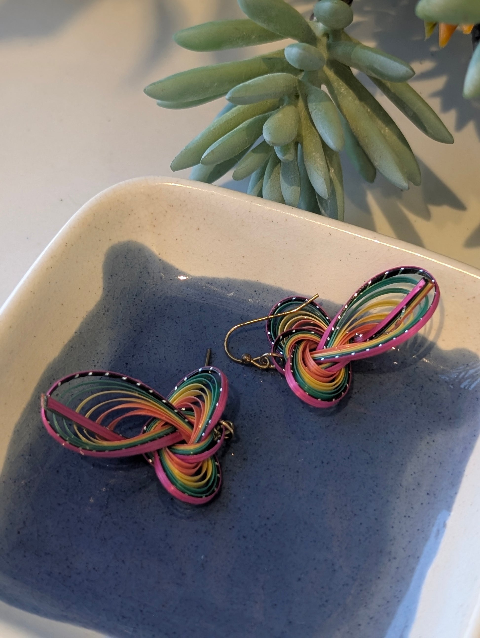 Colorful butterfly-shaped earrings on a textured surface with a plant in the background
